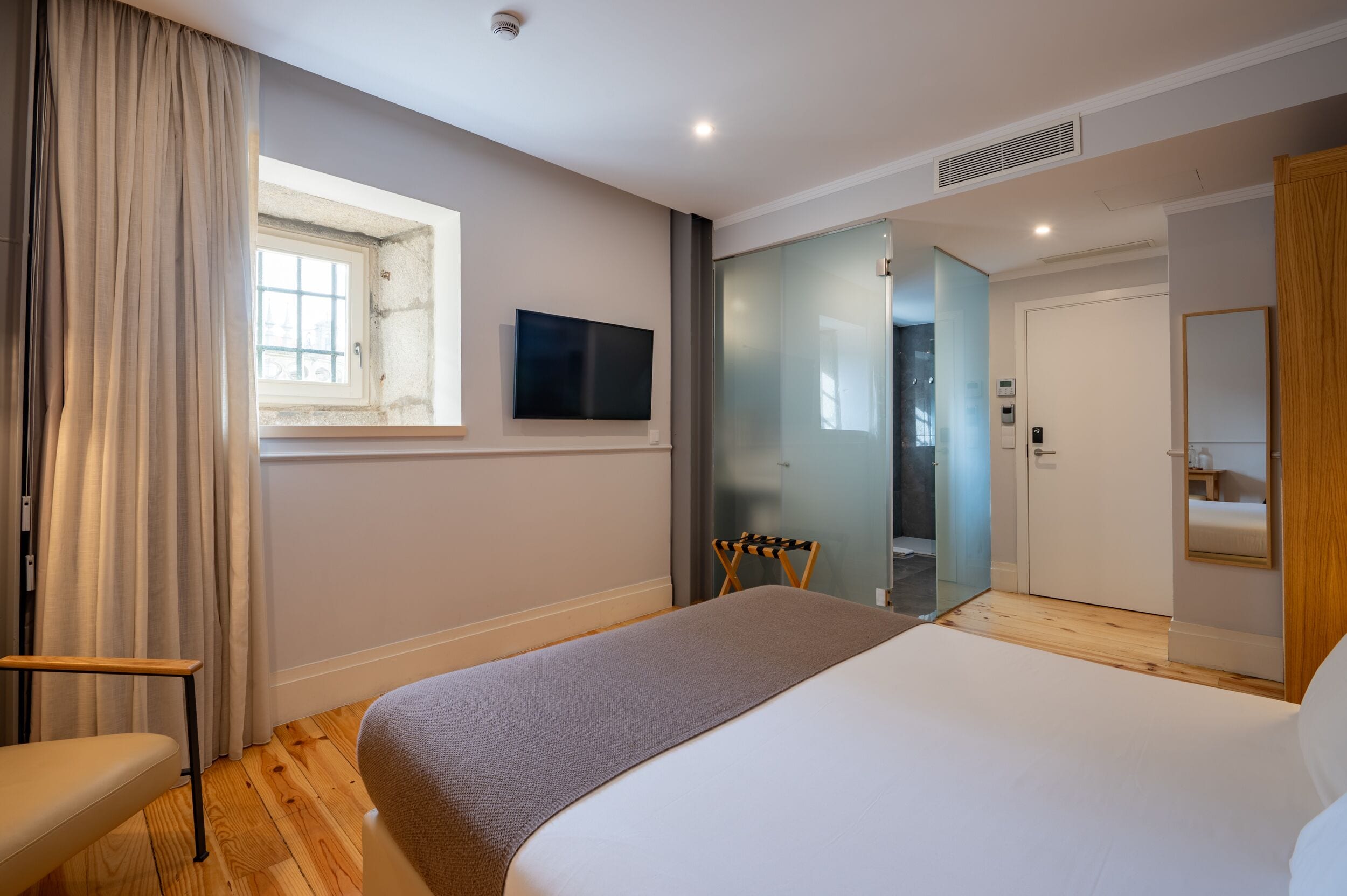 A modern hotel room with a large bed, a TV mounted on the wall, a window with curtains, a wooden floor, and a door leading to a bathroom.