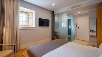 Una habitación de hotel moderna con una cama grande, un televisor fijado a la pared, una ventana con cortinas, suelo de madera y una puerta que da al baño.