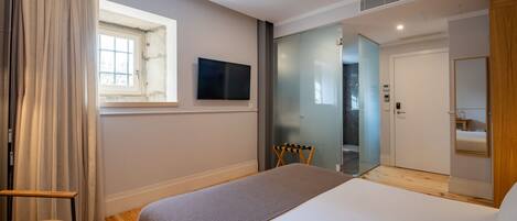 A modern hotel room with a large bed, a TV mounted on the wall, a window with curtains, a wooden floor, and a door leading to a bathroom.