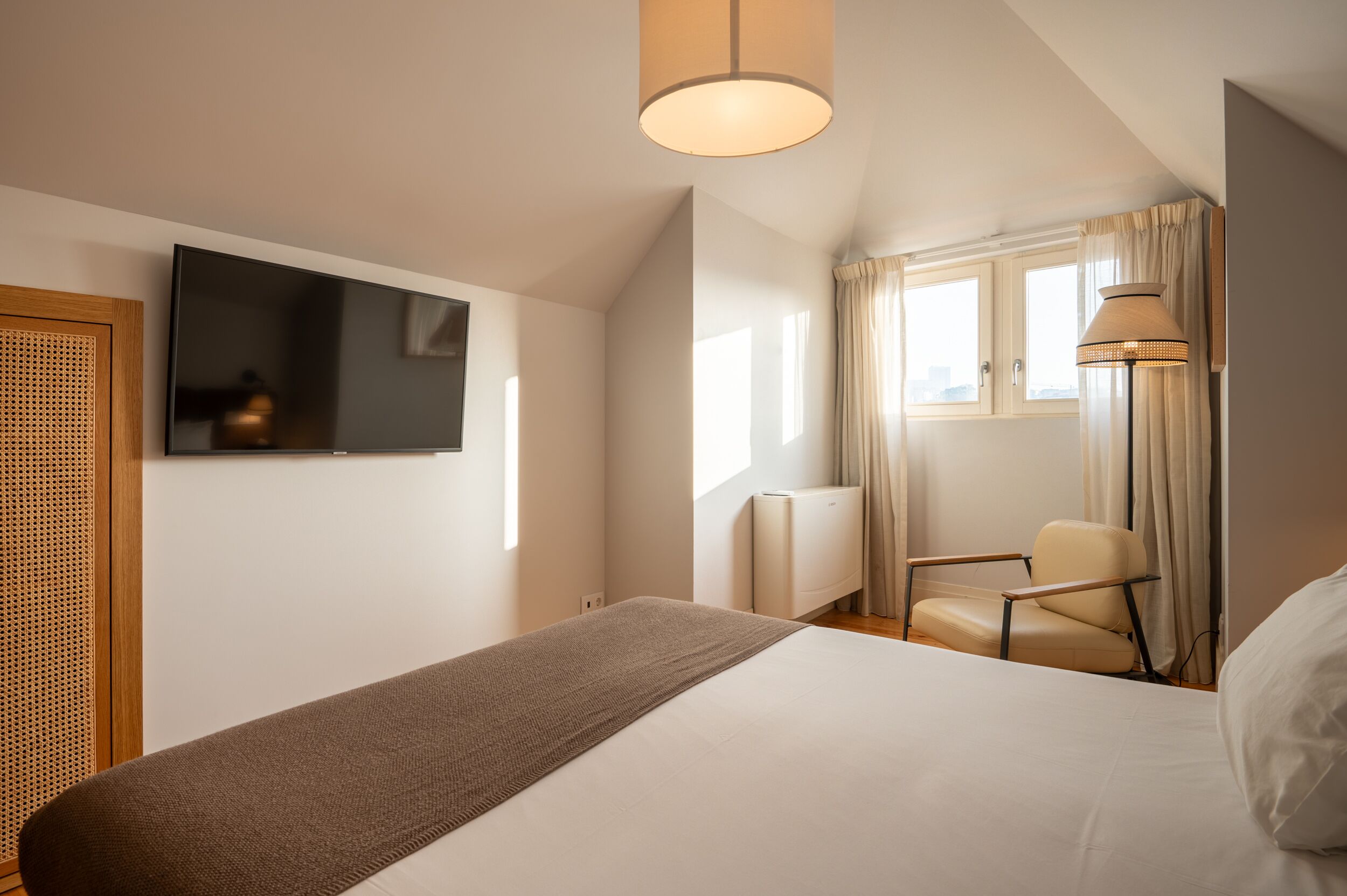 Habitación de hotel con cama, televisión montada en la pared, una silla, ventana con cortinas y una lámpara.