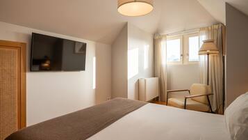 Habitación de hotel con cama, televisión montada en la pared, una silla, ventana con cortinas y una lámpara.