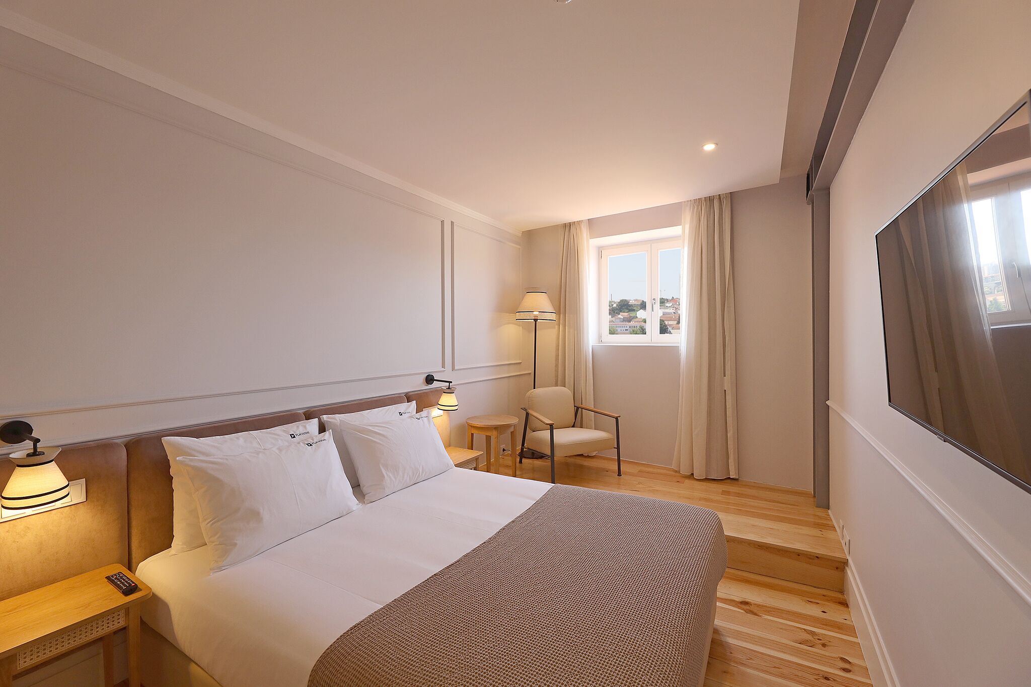 Habitación de hotel con cama, mesita de noche, lámpara, sillón y un amplio ventanal con cortinas.