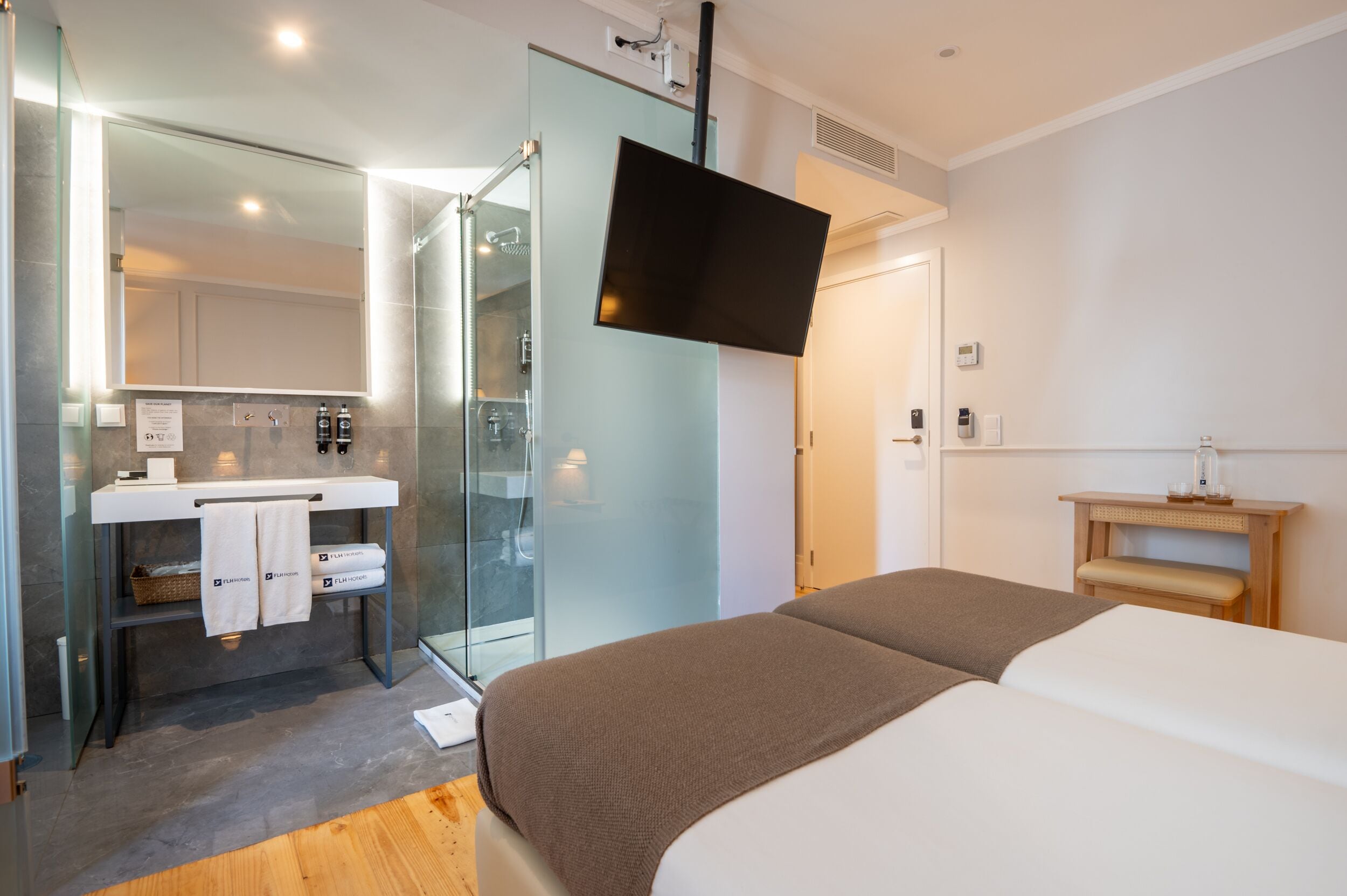Quarto de hotel moderno com chuveiro em cabine de vidro, televisão de tela plana na parede, mesa de cabeceira com luminária e cama com manta marrom.