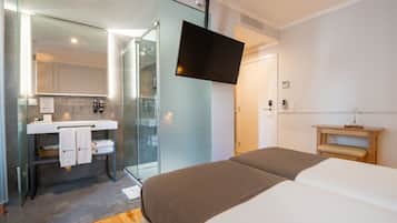 Quarto de hotel moderno com chuveiro em cabine de vidro, televisão de tela plana na parede, mesa de cabeceira com luminária e cama com manta marrom.