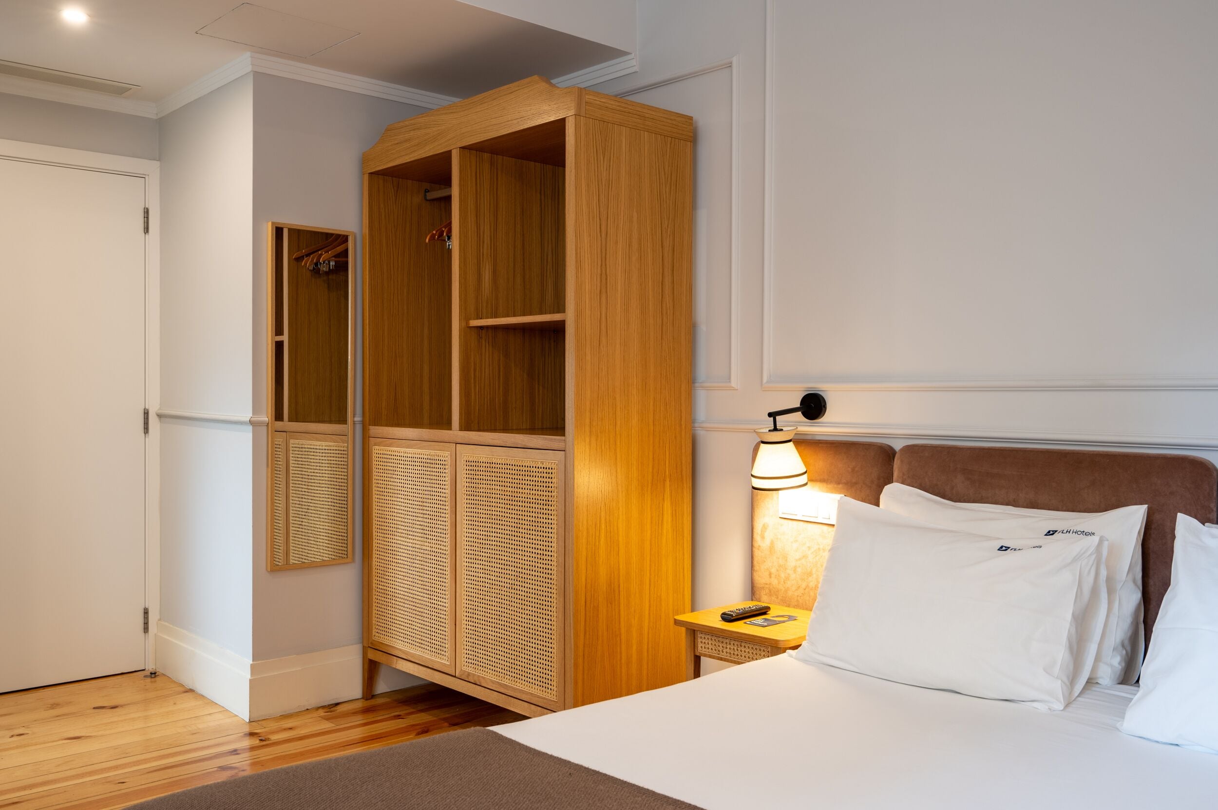 Een hotelkamer met een bed, een houten garderobe, een nachtkastje met een lamp en een spiegel.