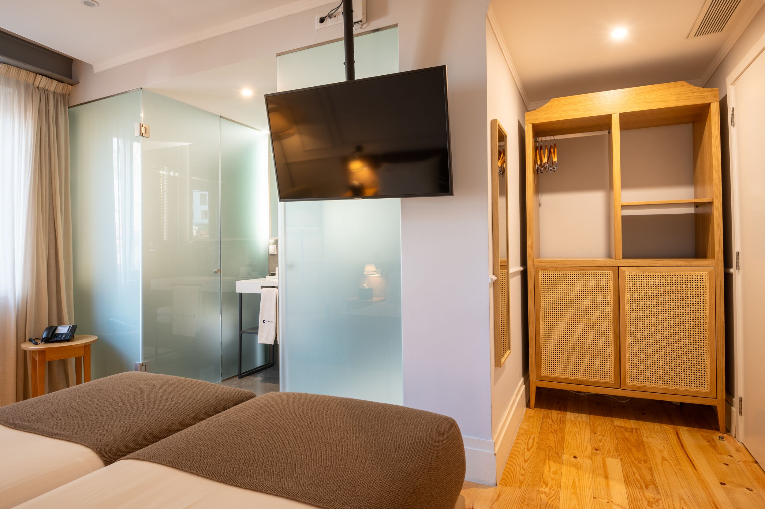 Habitación de hotel con cama, televisor montado en la pared, armario de madera y separador de vidrio que divide la habitación del baño.