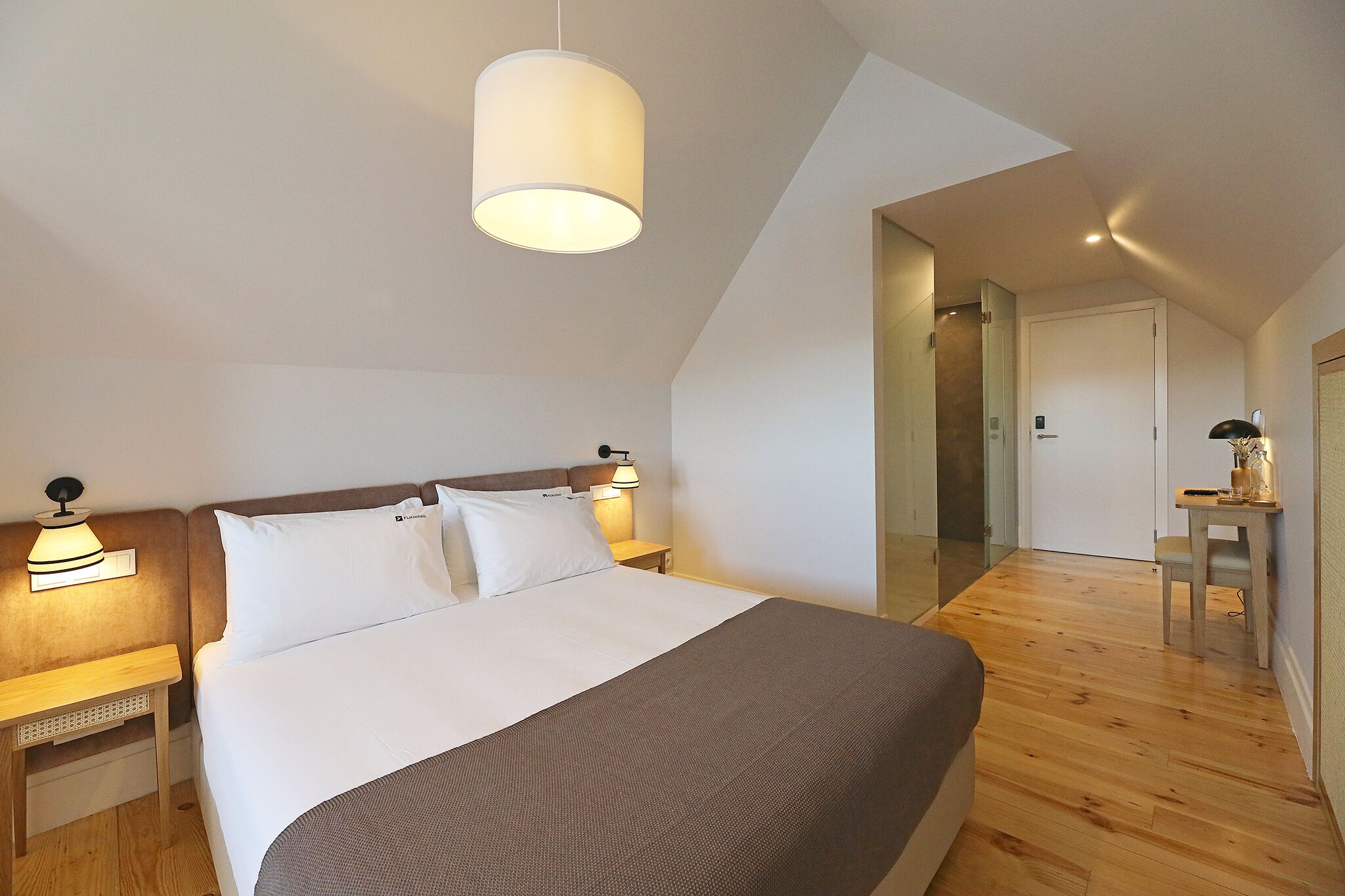 A modern hotel room with a large bed, a bedside table with a lamp, a small table with a lamp, and a mirror on the wall.
