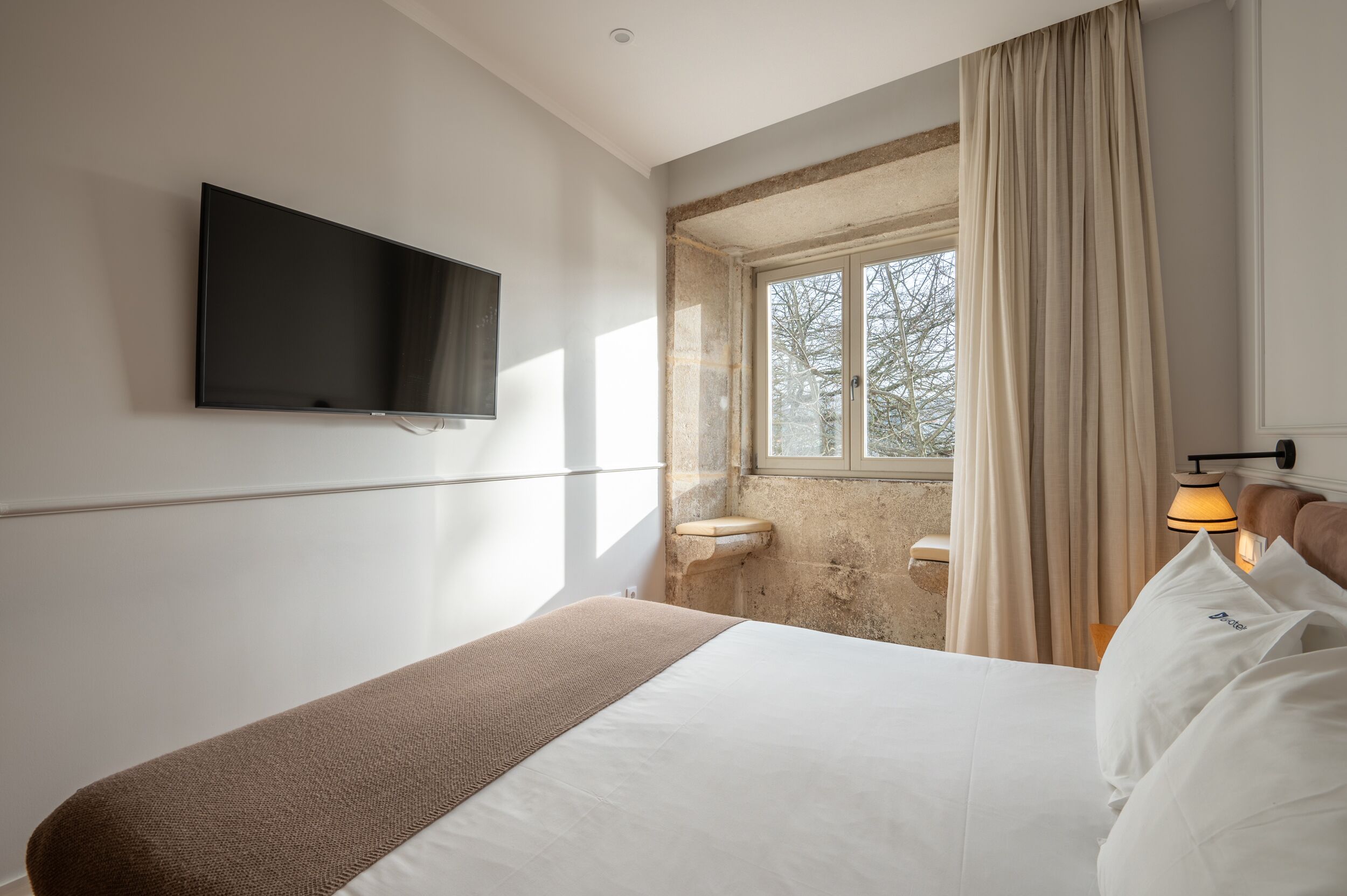 Quarto de hotel com uma cama, televisão, janela com cortinas e uma pia pequena.