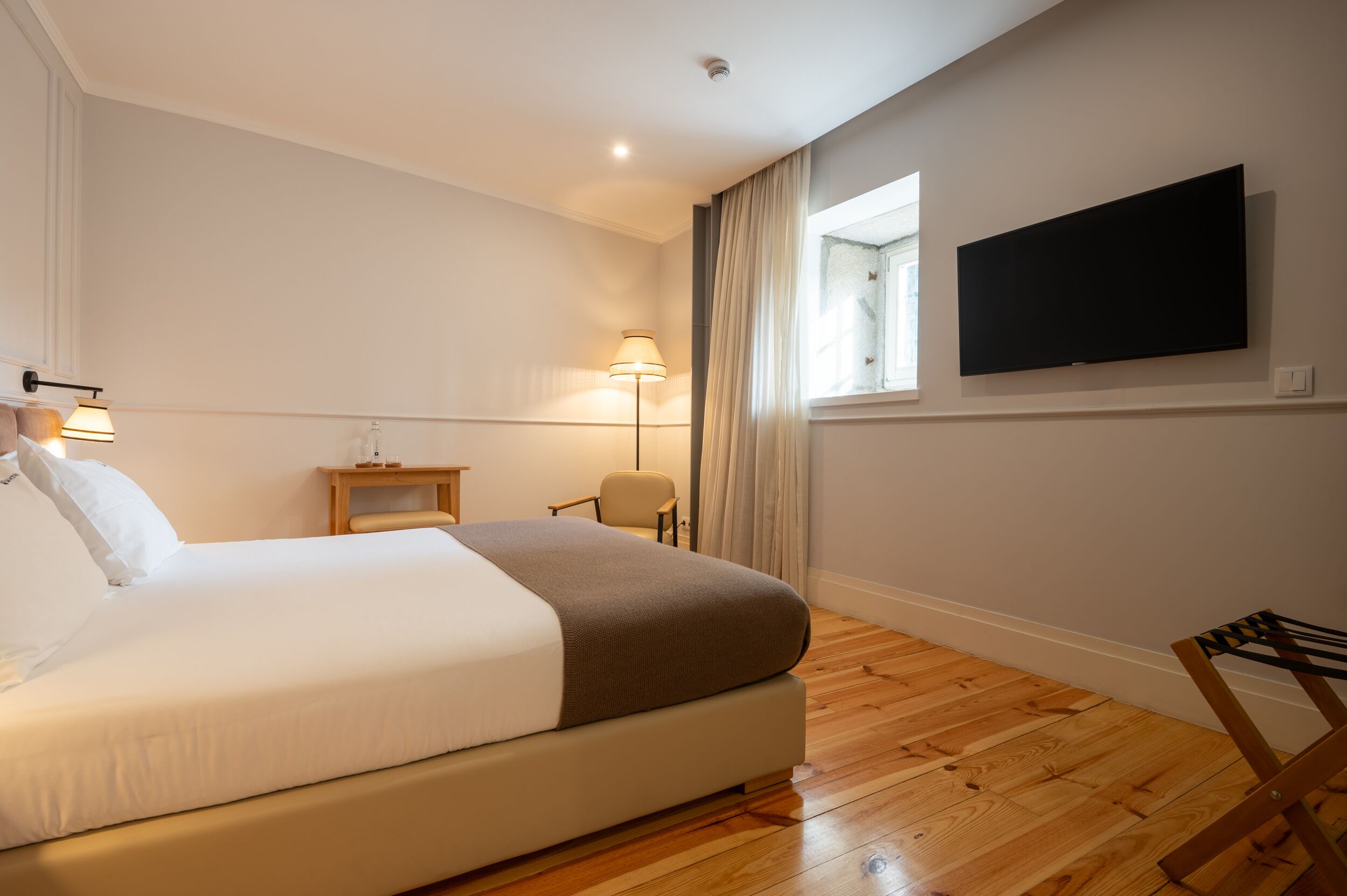 A hotel room with a large bed, a flat-screen TV mounted on the wall, a small bedside table, a chair, and a window with curtains.
