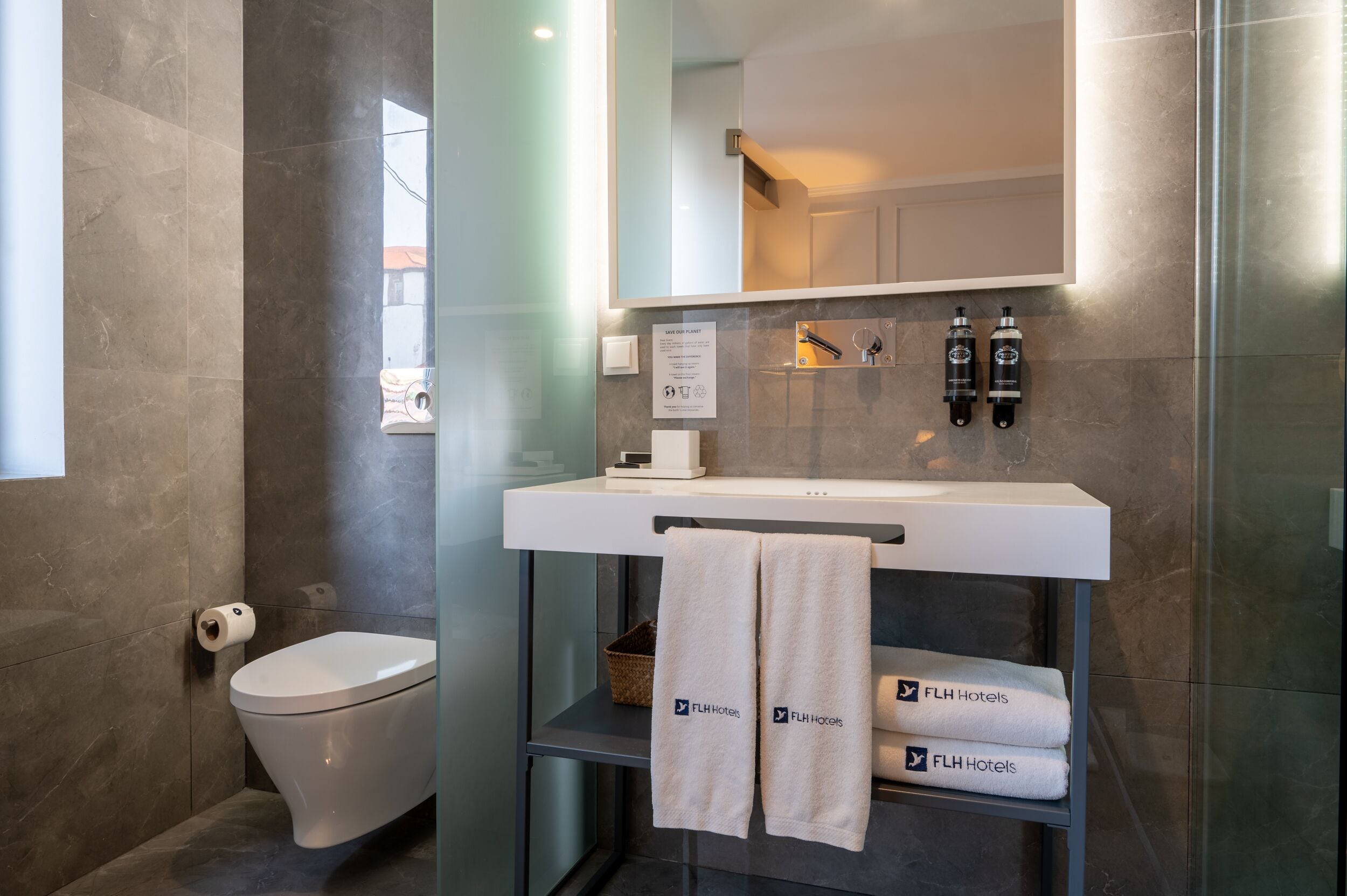 A modern bathroom with a toilet, a sink, and a mirror. There are towels hanging on a rack and a sign on the wall.