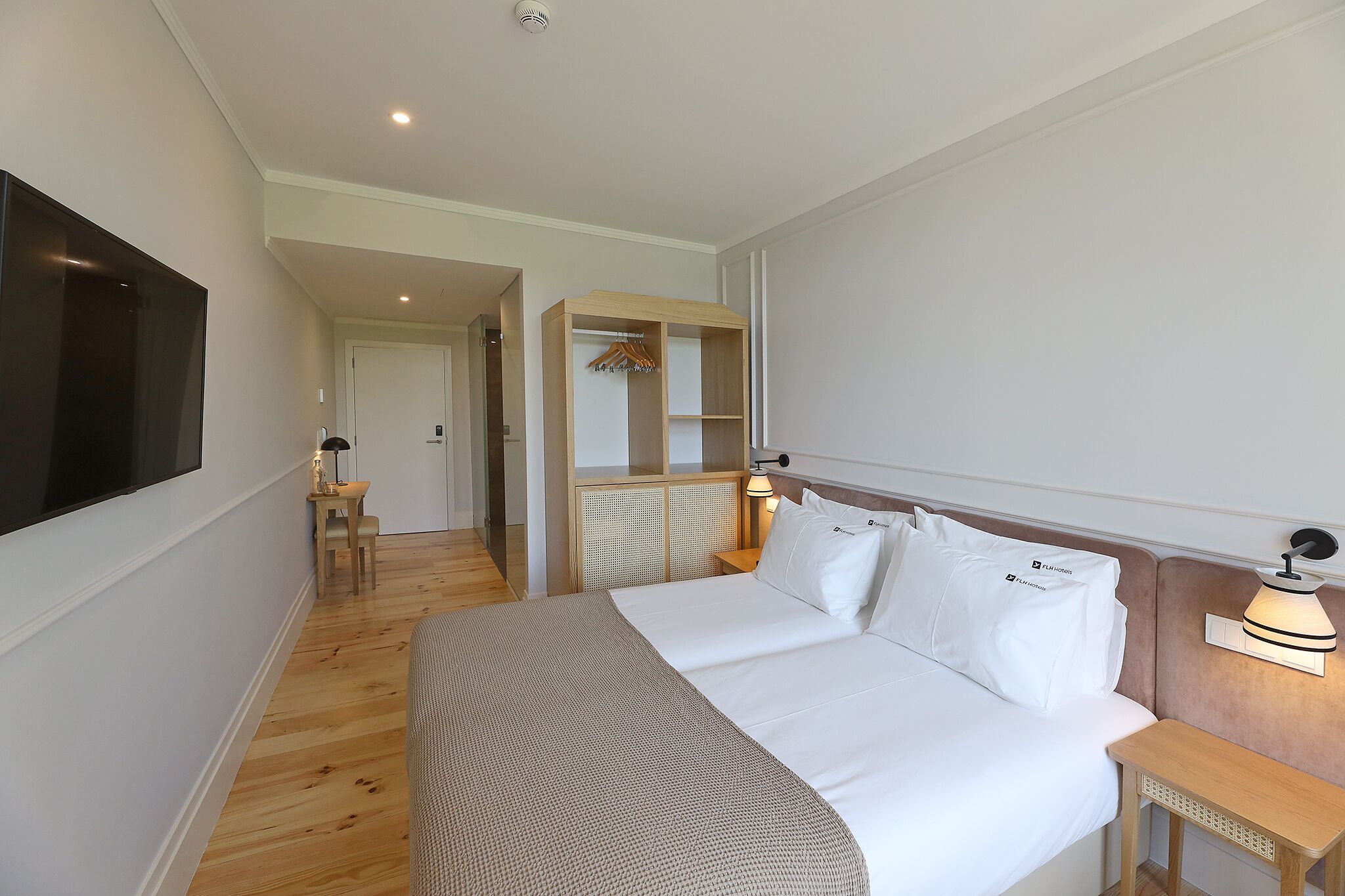 Una habitación de hotel con una cama grande, un televisor montado en la pared, una mesita de noche con una lámpara y un vestidor.
