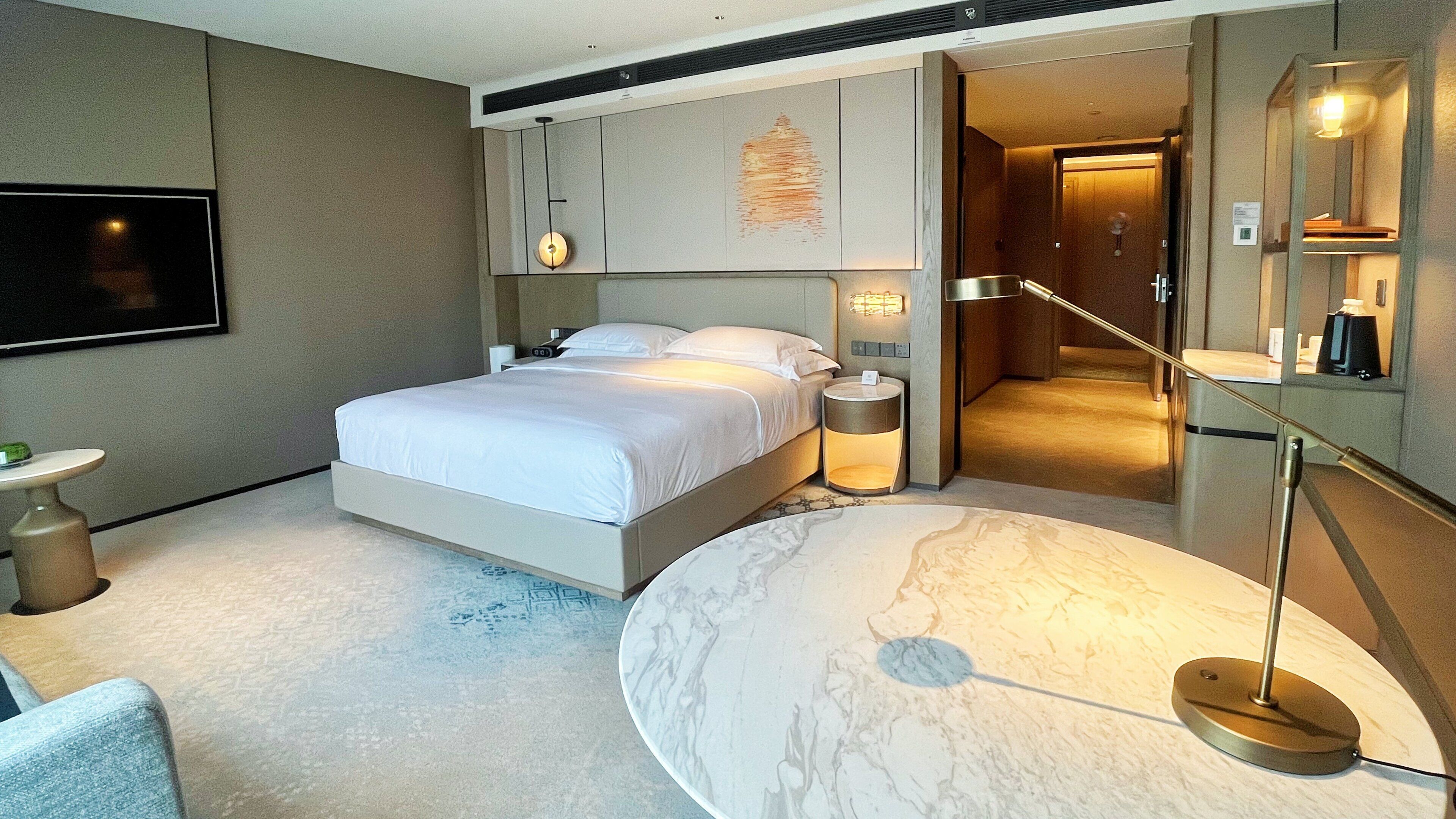 A modern hotel room with a large bed, bedside lamps, a marble accent table, and a flat-screen TV mounted on the wall.