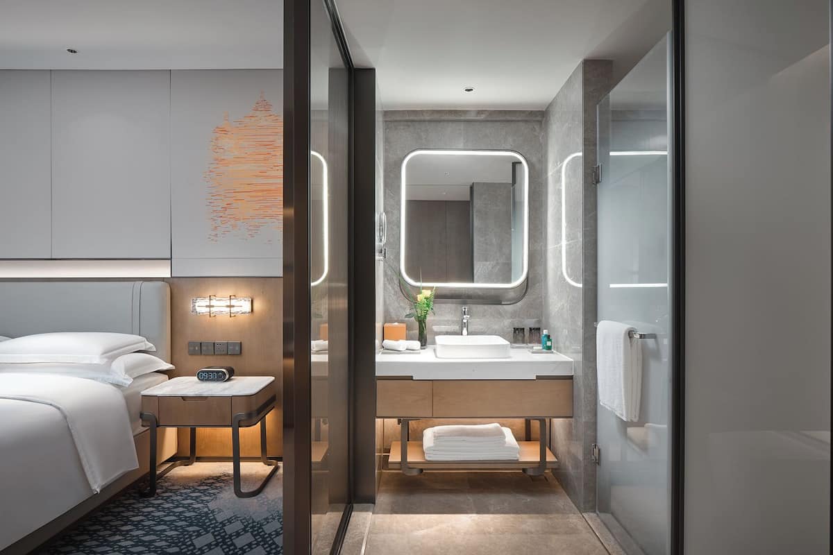 Una habitación de hotel moderna con cama, mesita de noche y baño con lavamanos, espejo y toallero.