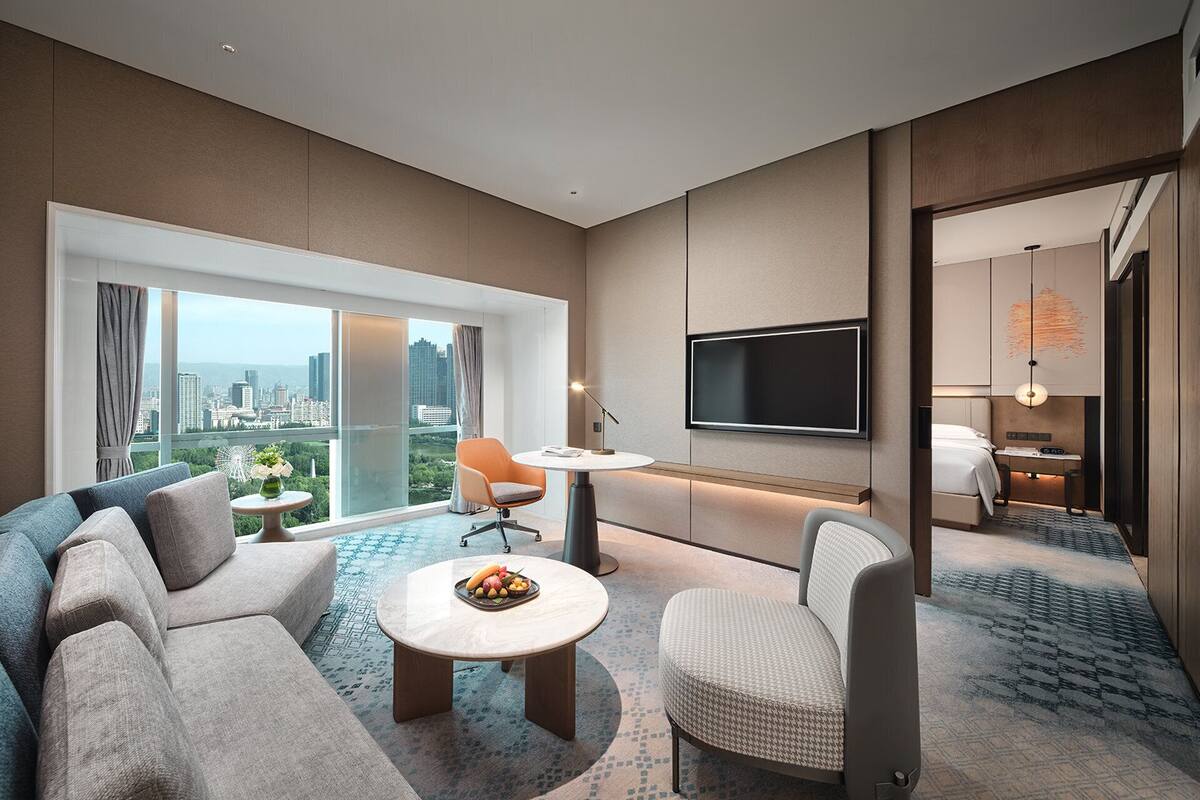 Habitación de hotel moderna con un amplio ventanal que ofrece vistas a la ciudad, un televisor de pantalla plana en la pared, un sofá, una mesita redonda con fruteros y una alfombra estampada.