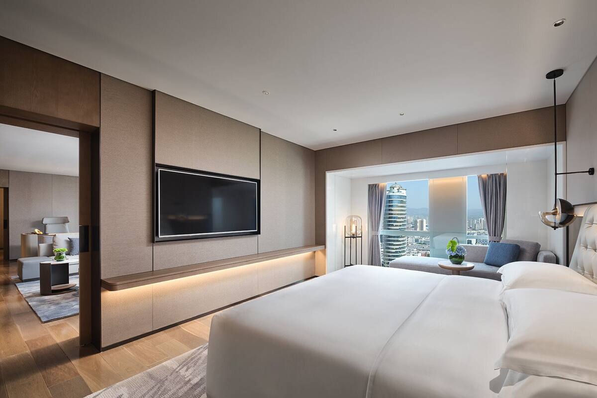 Una habitación de hotel moderna con una cama grande, un televisor de pantalla plana montado en la pared, vista a la ciudad por la ventana y un pequeño rincón de descanso con una mesa y sillas.