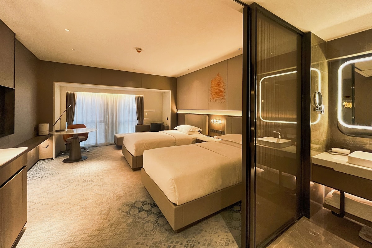 Habitación de hotel con dos camas, un escritorio, una silla, un ventanal grande con cortinas, un baño con espejo y lavamanos, y piso alfombrado.