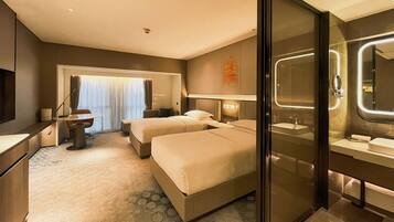A hotel room with two beds, a desk, a chair, a large window with curtains, a bathroom with a mirror and sink, and a carpeted floor.