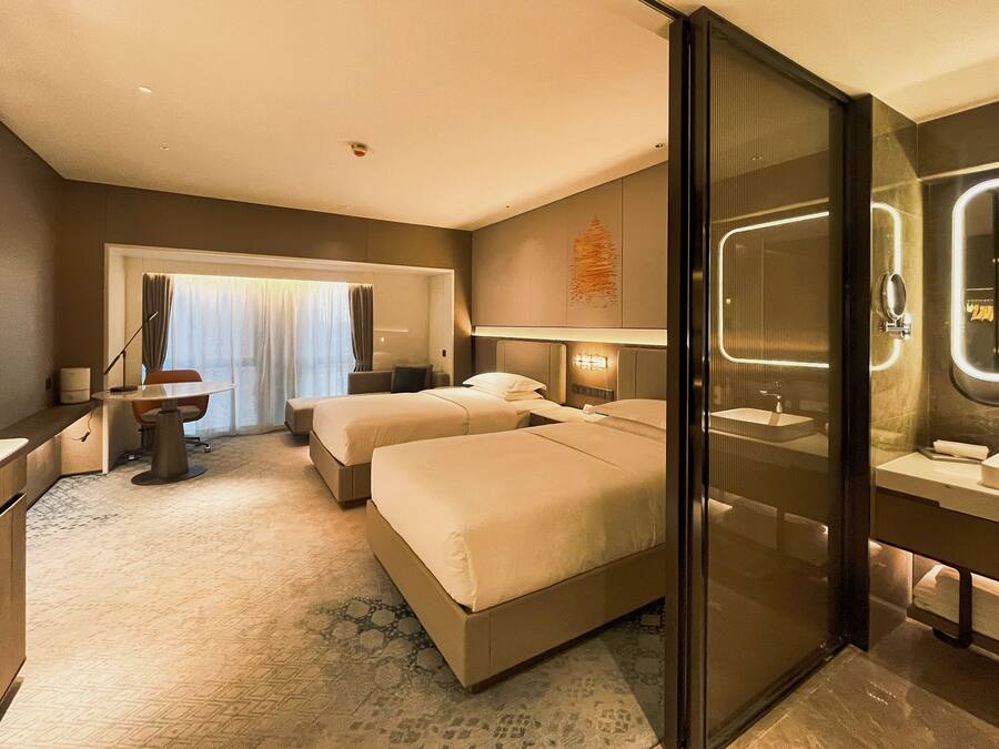 A hotel room with two beds, a desk, a chair, a large window with curtains, a bathroom with a mirror and sink, and a carpeted floor.