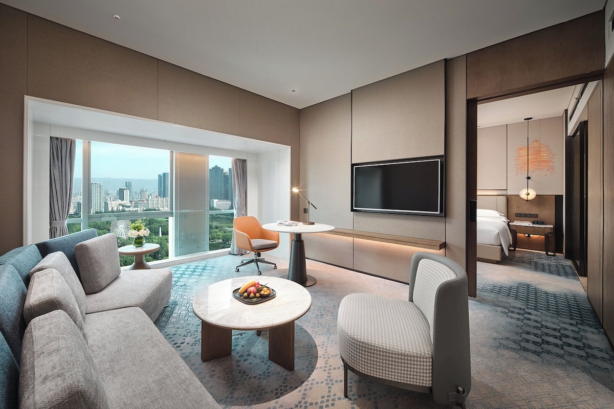 Habitación de hotel moderna con un amplio ventanal que ofrece vistas a la ciudad, un televisor de pantalla plana en la pared, un sofá, una mesita redonda con fruteros y una alfombra estampada.