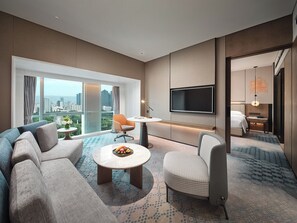 Una habitación de hotel moderna con un ventanal grande, un televisor de pantalla plana, un sofá, una silla y una mesita con fruta.