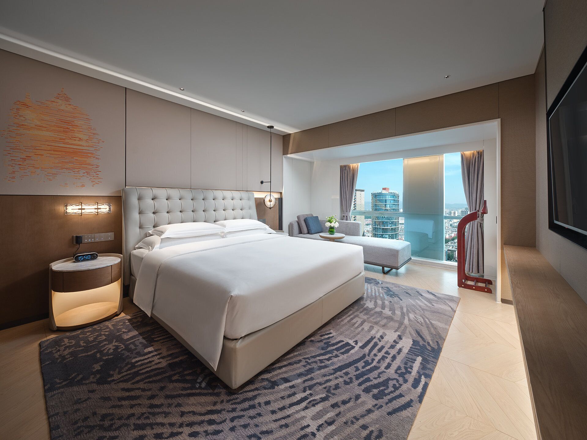 A modern hotel room with a large bed, a bedside table, a nightstand, a chair, a sofa, and a view of the city.