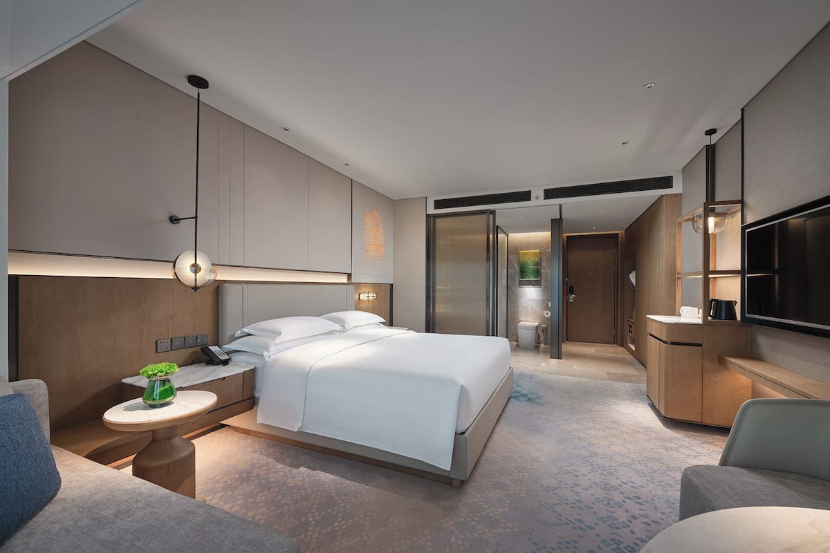 Una habitación de hotel moderna con una cama grande, mesitas de noche, un escritorio, un televisor y un baño visible a través de una puerta corrediza.