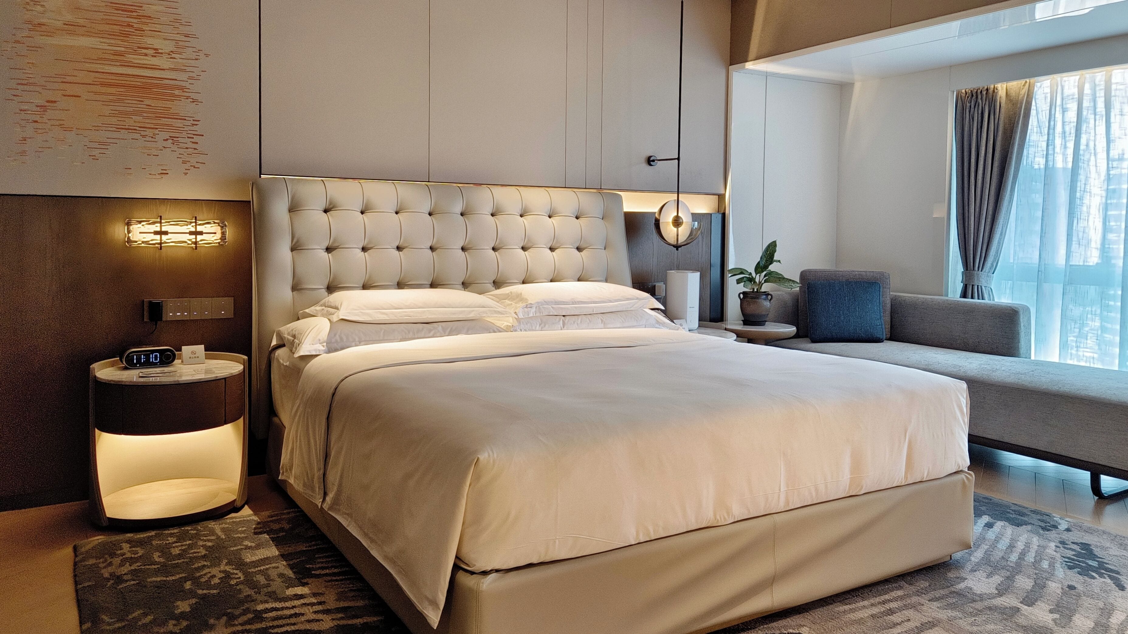 Una habitación de hotel moderna con una cama grande, una mesita de noche con un reloj digital, una lámpara de pared y una ventana con cortinas.