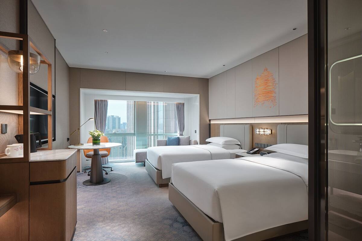 A modern hotel room with a desk, two beds, a large window, and a city view.