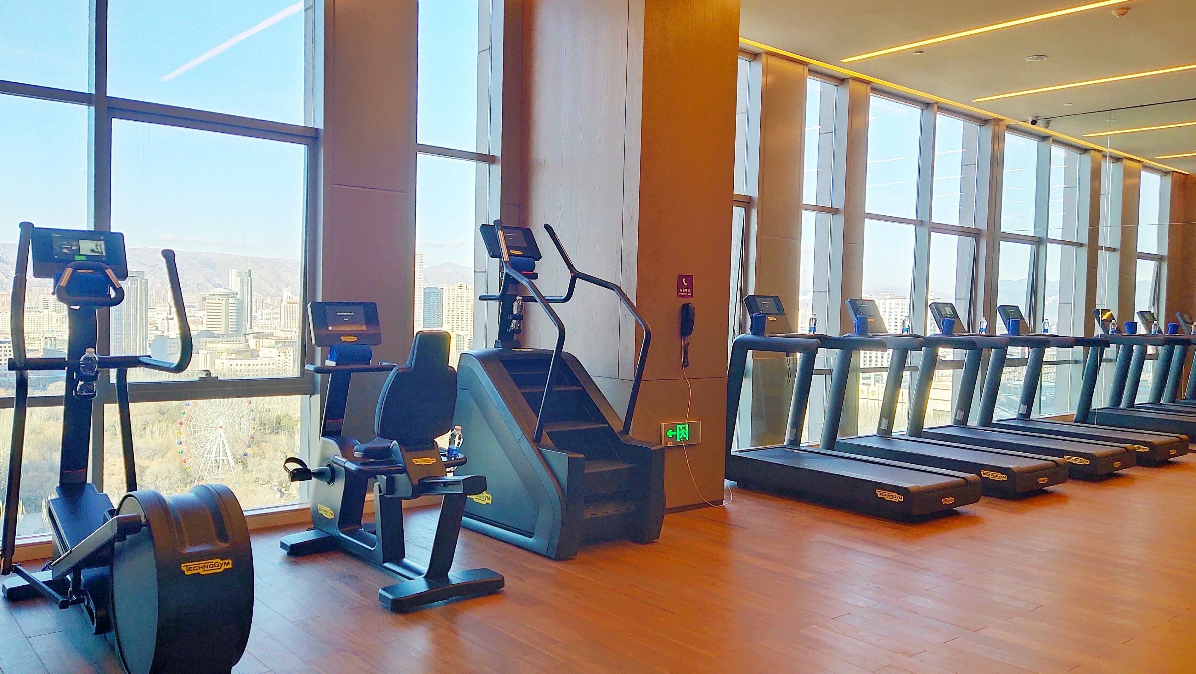 Een fitnessruimte met cardioapparaten en loopbanden, grote ramen en uitzicht op de stad.