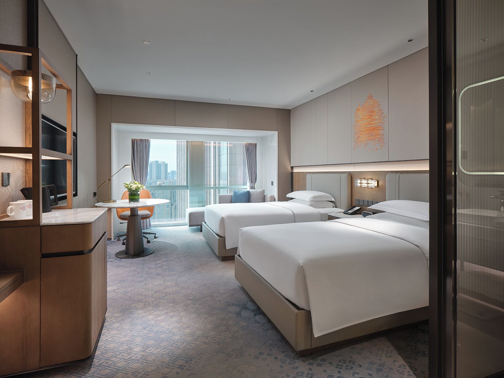 Een moderne hotelkamer met twee bedden, een bureau en een groot raam met uitzicht op de stad.