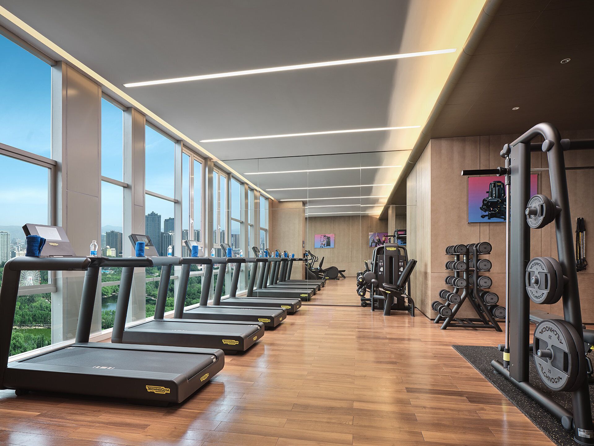 A gym with treadmills aligned along a wall with large windows, weight machines, and a wooden floor.