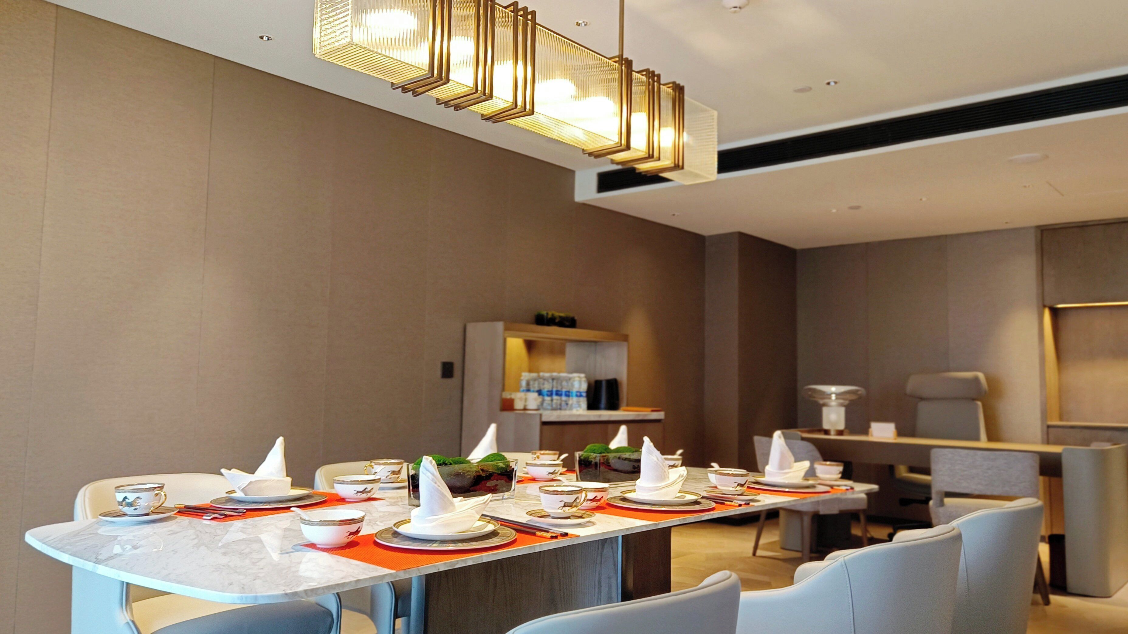 Un comedor moderno con una mesa larga dispuesta para una comida, sillas, una lámpara decorativa y un estante con agua embotellada.