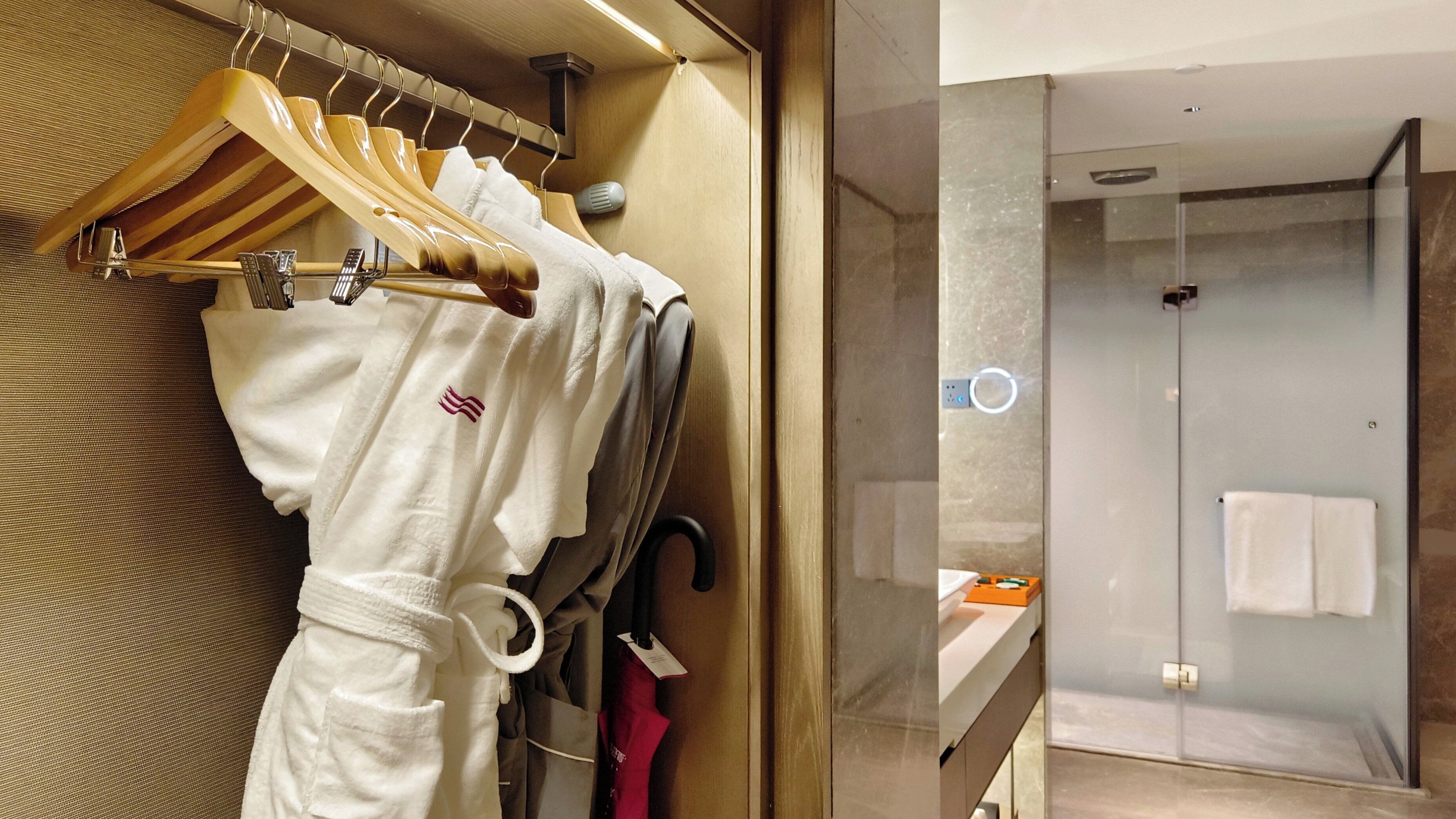 Un baño de hotel con cabina de ducha de vidrio, un perchero con varias toallas y una percha de madera con bata.
