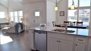 A spacious kitchen with a large island, white cabinetry, and a dishwasher. There is a dining area with a table and chairs, and a living room with a sofa and a lamp. - Bozeman Southside Retreat- 30+ Night Rental Home on Bozeman's South Side (Bozeman)
