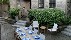 A wooden patio table set for six with white chairs, a stone pathway, and a wooden fence in the background.