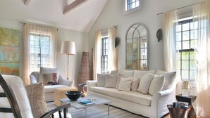 A spacious living room with a large sofa, a coffee table, and a chair. There are multiple windows providing natural light and a view of greenery outside.