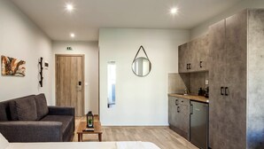 Una habitación moderna con un sofá gris, una mesita de madera con un farol, una cama, un espejo en la pared y una kitchenette con armarios y un refrigerador.