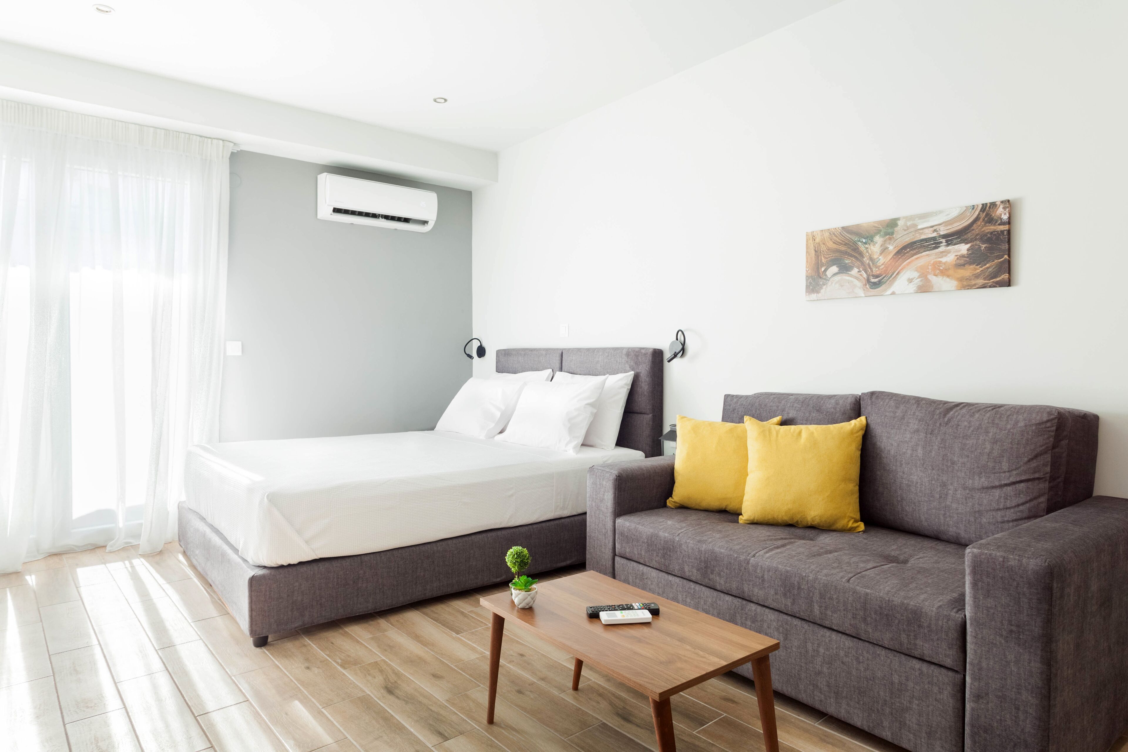 A modern room with a bed, a gray sofa with yellow pillows, a small table, and a painting on the wall.
