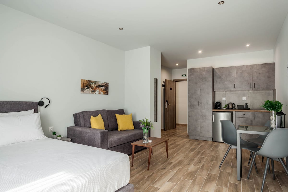 Une chambre d’hôtel moderne comprenant un lit, un canapé gris avec des coussins jaunes, une petite table avec un vase, et un coin cuisine équipé d’un réfrigérateur, d’une cuisinière et d’un évier.
