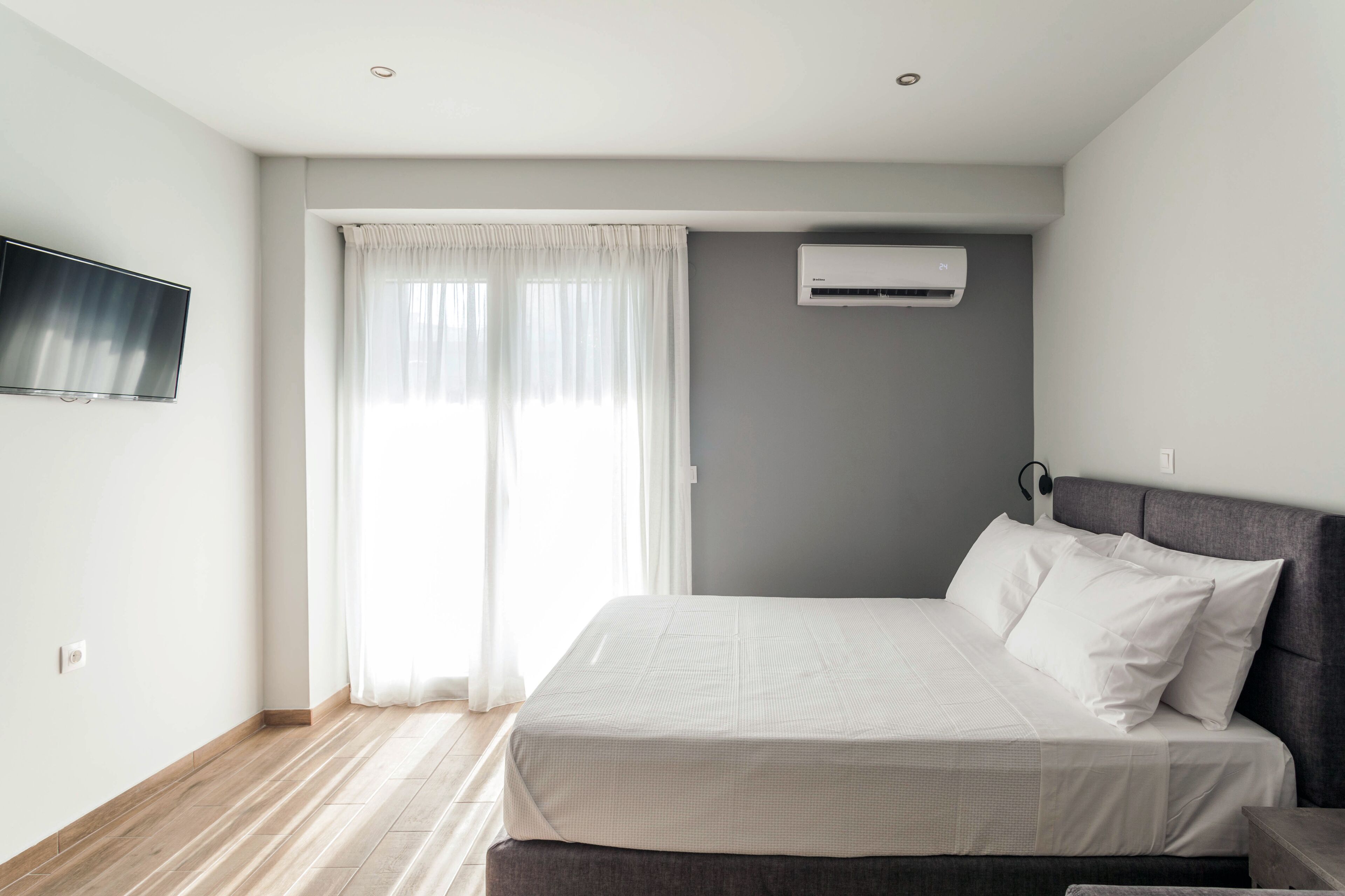 A modern bedroom with a bed, a flat-screen TV, an air conditioner, and a window with curtains.