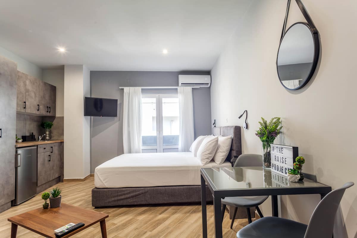 Une chambre moderne avec un lit, une table de chevet et un petit bureau avec une chaise. Un téléviseur est fixé au mur, il y a une fenêtre avec des rideaux et un appareil de climatisation.