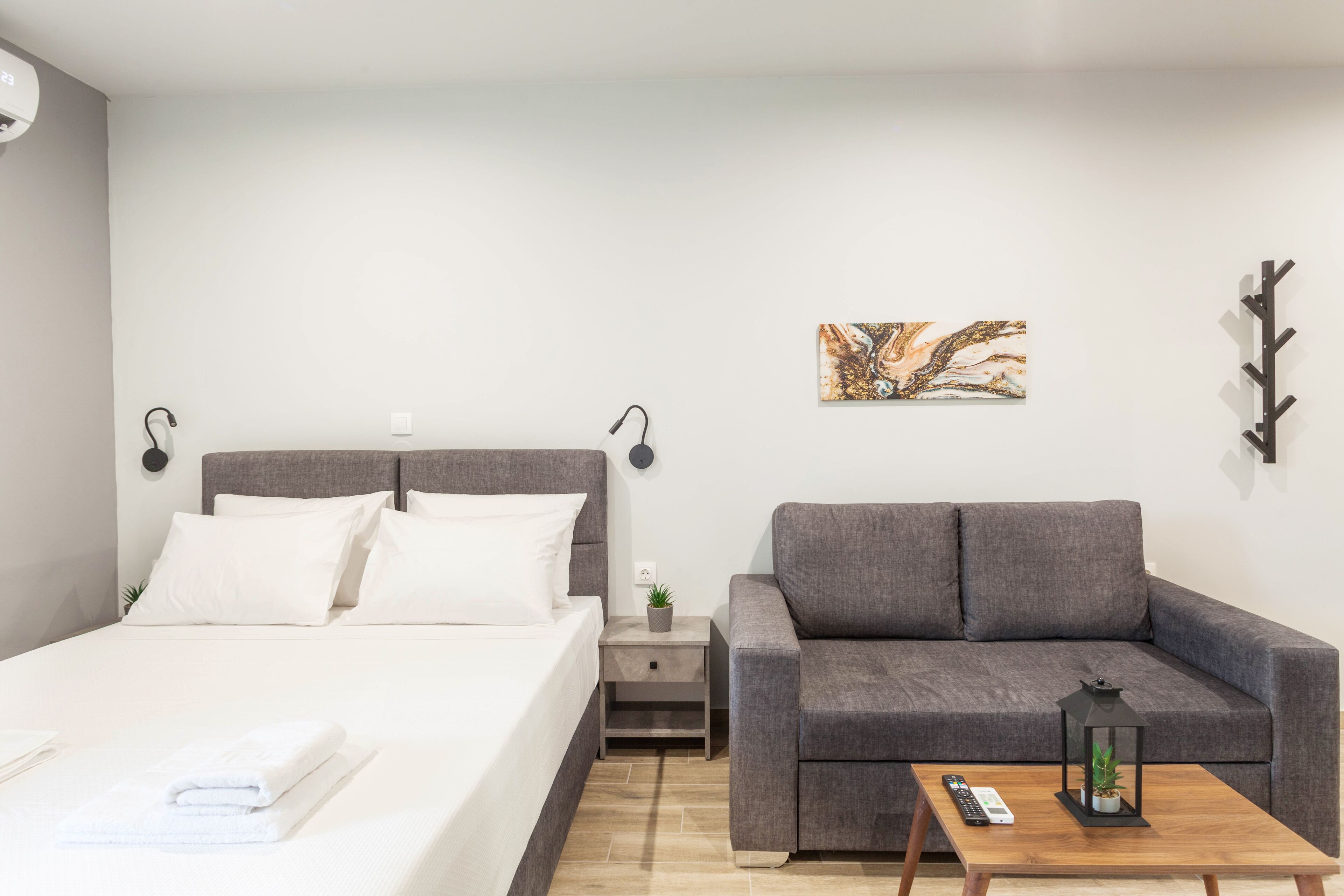 A modern hotel room with a bed, a gray sofa, a wooden coffee table, and a nightstand with a lamp and a plant.