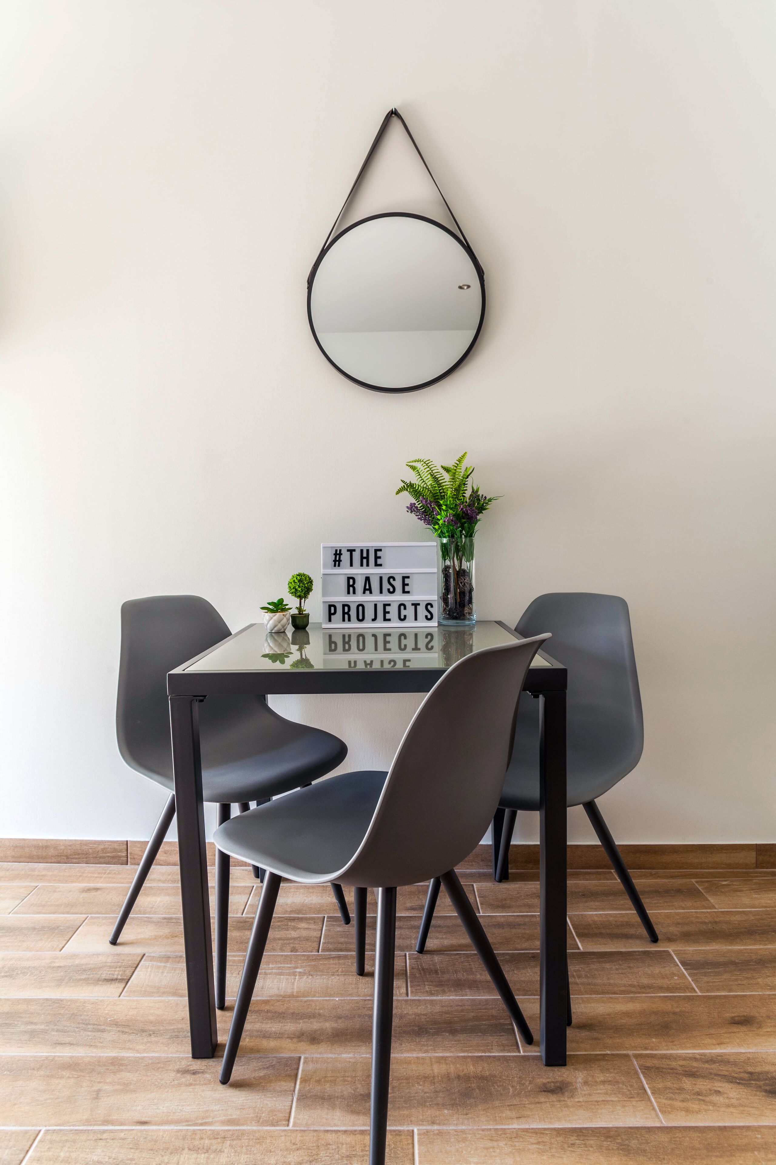 A modern dining area with a glass table, four grey chairs, a decorative mirror, a sign with motivational text, and a small plant.