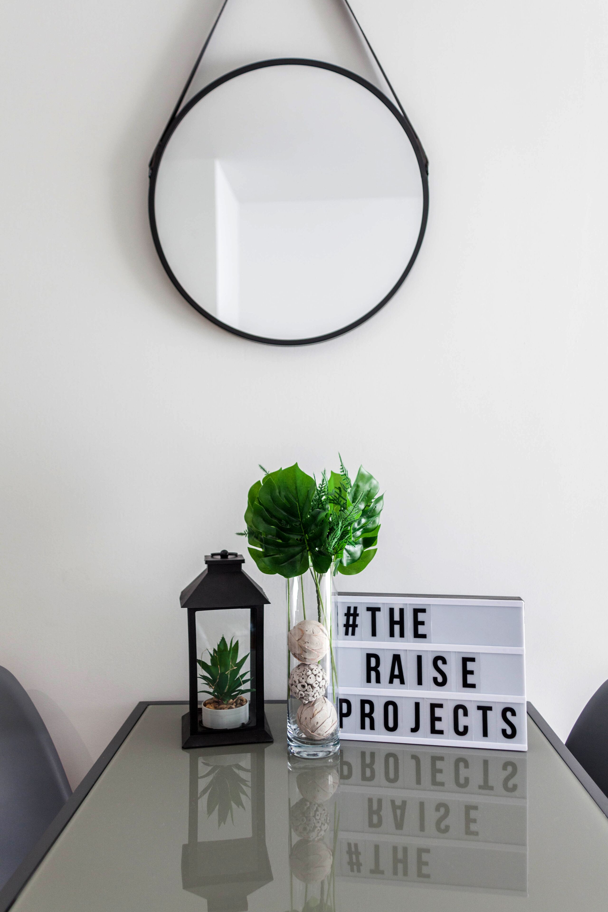 Une table avec un plateau en verre, une petite plante en pot, une lanterne et un panneau lumineux avec le texte « #THE RAISE PROJECTS ».