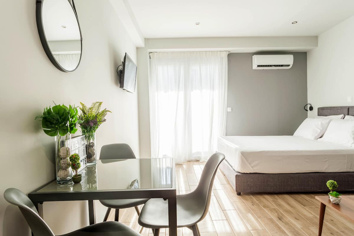 Une chambre d’hôtel moderne avec une table en verre, deux chaises, un lit avec une tête de lit grise, un appareil de climatisation et une grande fenêtre avec des rideaux légers.