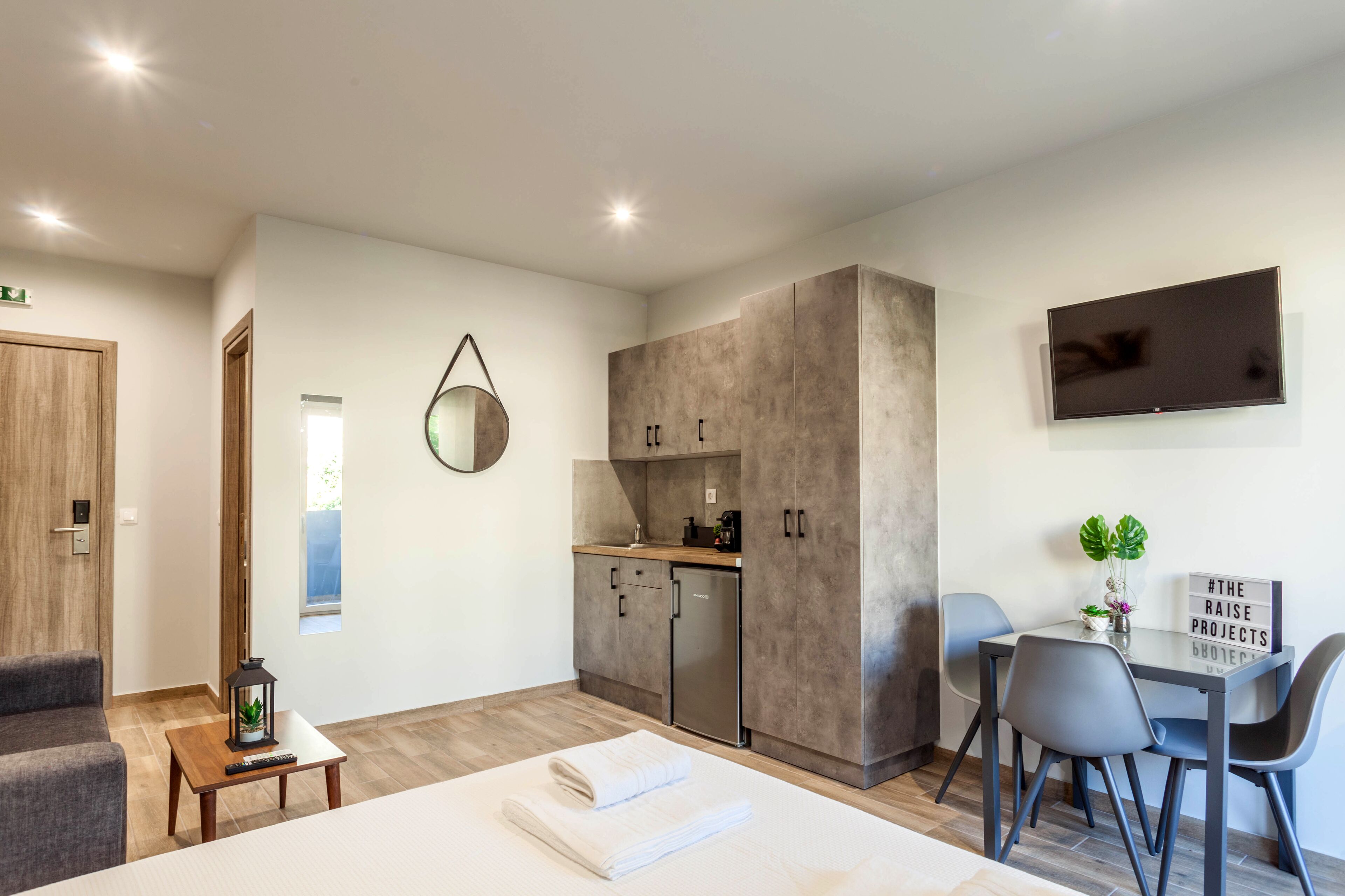 Una habitación de hotel moderna con cama, escritorio y silla, una mesita con planta, televisor montado en la pared y una zona de kitchenette.