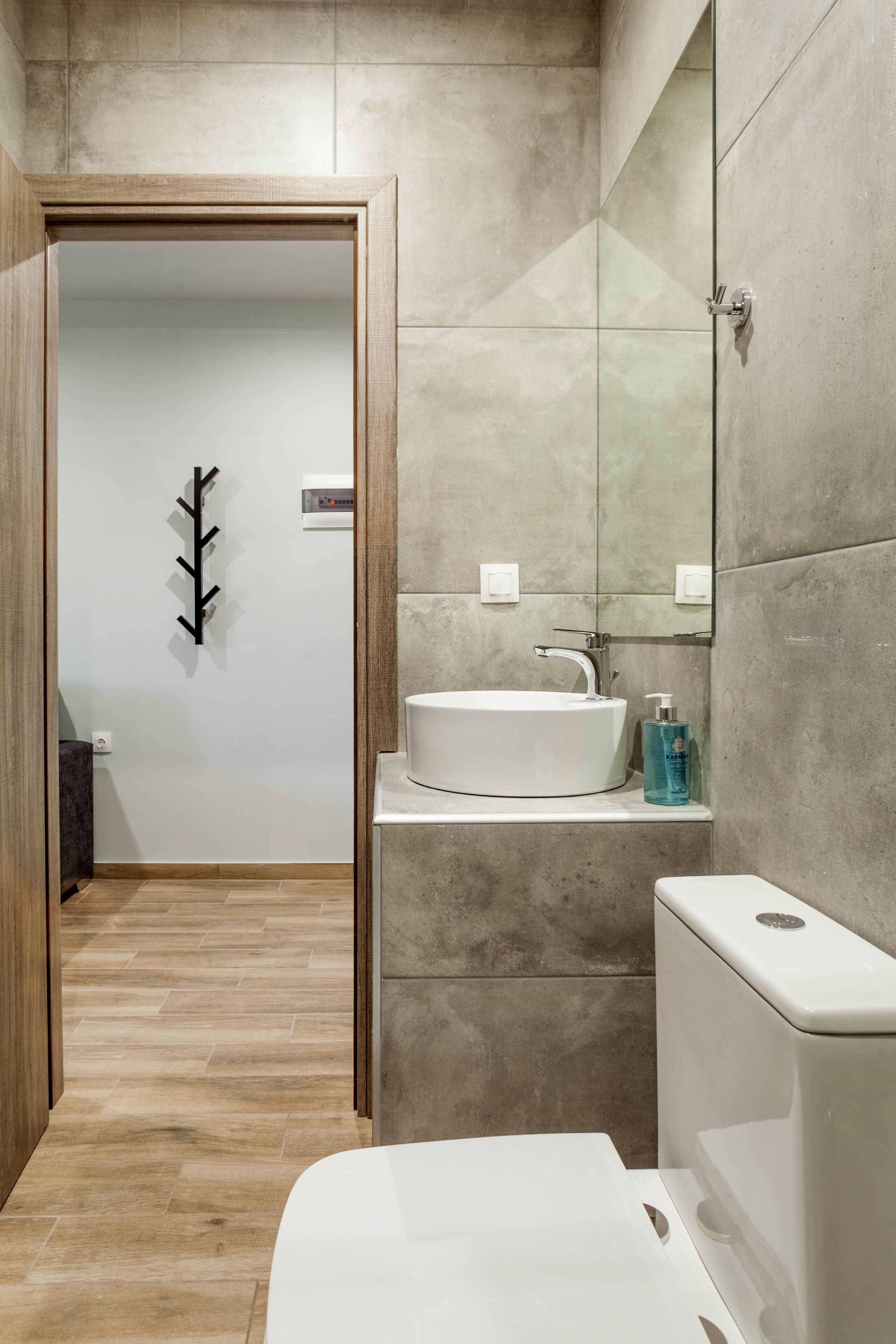 Un baño moderno con un lavamanos blanco, un espejo y un inodoro. Se observa una puerta de madera que da a otra habitación.