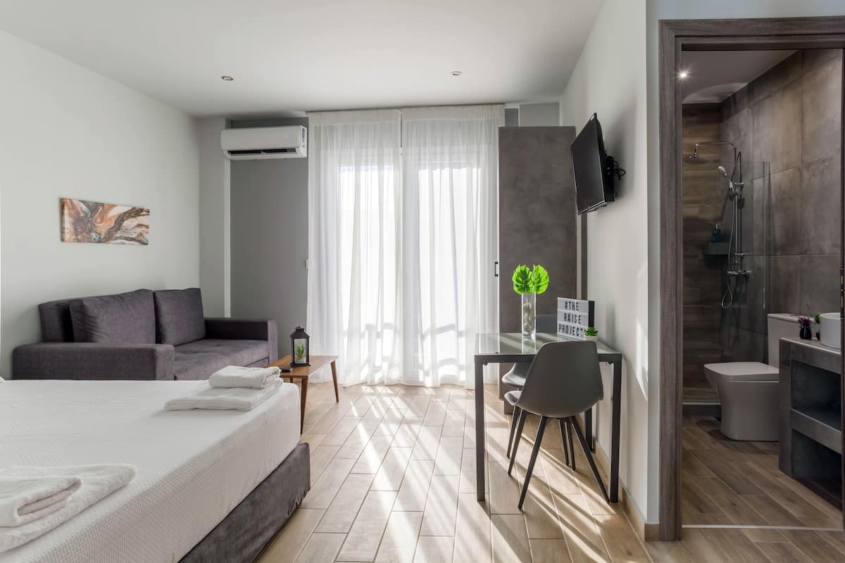 Une chambre d’hôtel moderne avec un lit, un canapé gris, un coin repas et une douche.