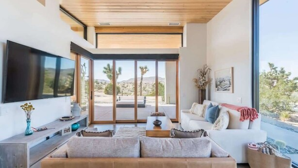 A modern living room with a flat-screen TV, a sofa, a coffee table, and a large window offering a view of palm trees and mountains. - Skyline Ridge By Homestead Modern (Yucca Valley)