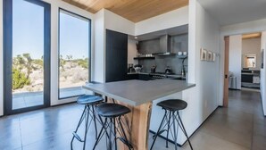 A modern kitchen with large windows, a central island, and bar stools. - Skyline Ridge By Homestead Modern (Yucca Valley)